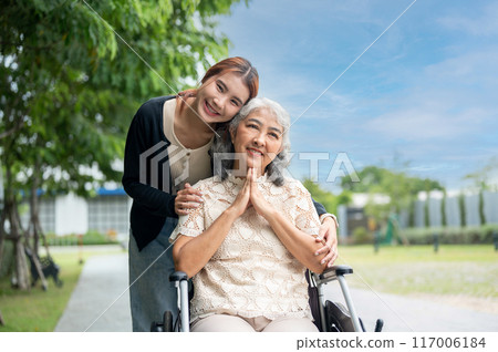 A lovely granddaughter is hugging her grandma, who is in a wheelchair, while walking her in a park. 117006184