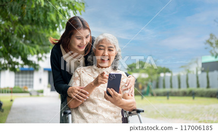 A lovely Asian granddaughter is hugging her grandma who is using a smartphone in a wheelchair. 117006185