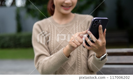 A close-up image of a happy Asian woman is using her smartphone on a bench in a garden. 117006202