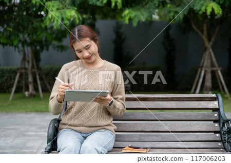 A beautiful Asian woman using her digital tablet on a bench in a park, working remotely. 117006203