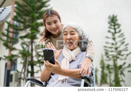 A happy Asian grandma in wheelchair and her granddaughter are enjoying a video call with a relative. 117006227