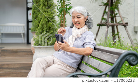 A positive, happy retired Asian woman is using her smartphone while relaxing in her home backyard. 117006228