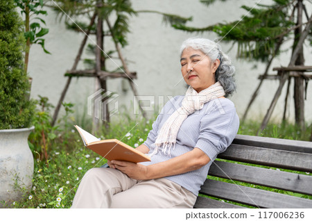 A calm mature Asian woman is sitting on a bench, her eyes closed and deep in thought while reading. 117006236