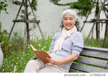 A happy retired Asian woman is smiling at the camera while sitting on a bench, holding a book. 117006238