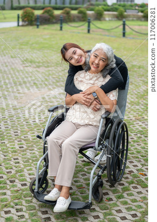 A lovely granddaughter is hugging her grandma, who is in a wheelchair, while walking her in a park. A lovely granddaughter is hugging her grandma, who is in a wheelchair, while walking her in a park. 117006281