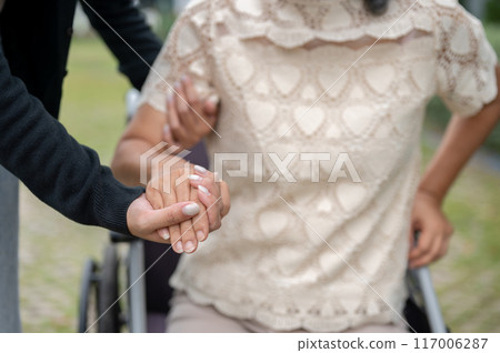 A caring granddaughter holding her grandma's hand, helping her stand up from the wheelchair. A caring granddaughter holding her grandma's hand, helping her stand up from the wheelchair. 117006287