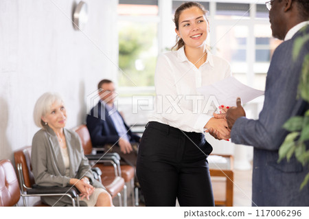 Woman makes deal, shaking hands with middle-aged African man colleague while standing in office 117006296