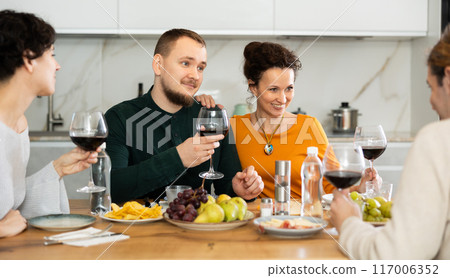 Group of joyful individuals are celebrating a holiday, enjoying drinks and conversation around table in their home Group of joyful individuals are celebrating a holiday, enjoying drinks and conversation around table in their home 117006352