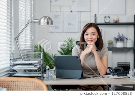 A young woman working from home at her desk with a tablet and laptop, smiling confidently. Modern home office setup with natural light. A young woman working from home at her desk with a tablet and laptop, smiling confidently. Modern home office setup with natural light. 117008838