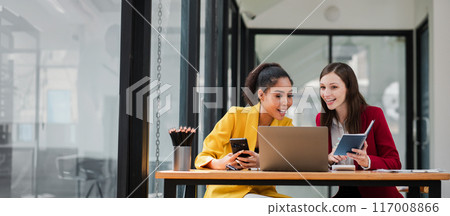Two professional women working together in a modern office, using laptops and notebooks, smiling and discussing ideas. 117008866