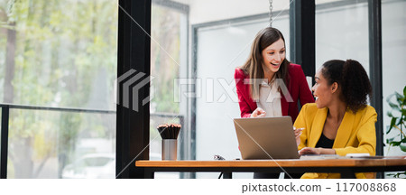 Two professional women working together on a laptop in a contemporary office setting with large windows and natural light. 117008868