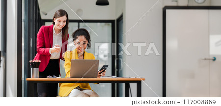 Two professional women working together in a modern office, using a laptop and smartphone, showcasing teamwork and productivity. Two professional women working together in a modern office, using a laptop and smartphone, showcasing teamwork and productivity. 117008869