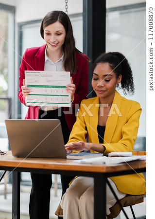 Two professional women working together on a financial report in a contemporary office environment, showcasing teamwork and productivity. 117008870