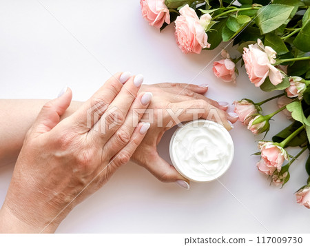 Hands middle age woman hold an open jar of white hand or body cream next to pink tea roses on white background, top view. Flat lay composition. Skincare and beauty product concept. Hands middle age woman hold an open jar of white hand or body cream next to pink tea roses on white background, top view. Flat lay composition. Skincare and beauty product concept. 117009730