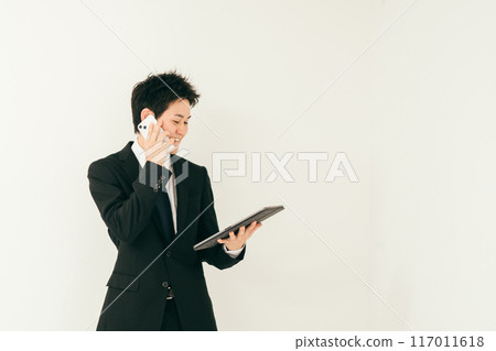 A man in his 20s in a suit talking on the phone while looking at documents 117011618