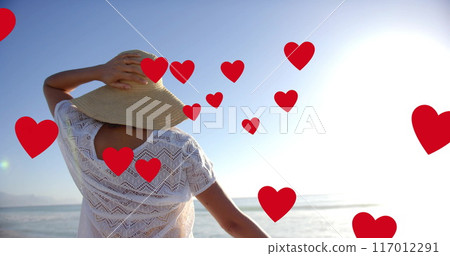 Image of hearts moving over biracial woman in hat on beach in summer 117012291