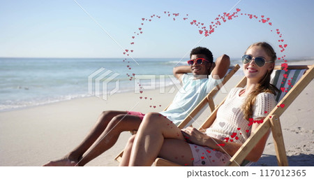 Image of hearts moving over diverse couple in love in deckchairs on beach in summer 117012365
