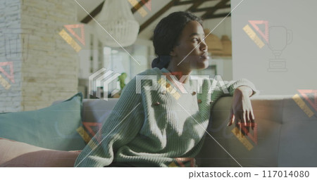 The image shows a thoughtful african american woman sitting on a sofa and looking outside a window The image shows a thoughtful african american woman sitting on a sofa and looking outside a window 117014080