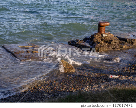 Old rusty bollard on the sea. Batumi. The sea is overflowing the pier. Old concrete slabs on the shore. Fortified coast. 117017471