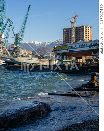 Ships and cranes in the cargo port. Old rusty bollard on the sea. Batumi. The sea overwhelms the pier. Old concrete slabs on the shore. Fortified coast. Black Sea Ships and cranes in the cargo port. Old rusty bollard on the sea. Batumi. The sea overwhelms the pier. Old concrete slabs on the shore. Fortified coast. Black Sea 117017489