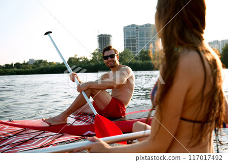 Young man and woman in swimwear sitting on sup boards, sunbathing, talking, relaxing on river. Paddle boarding activity Young man and woman in swimwear sitting on sup boards, sunbathing, talking, relaxing on river. Paddle boarding activity 117017492
