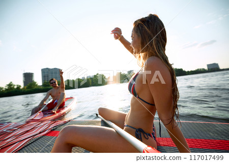 Young man and woman in swimwear sitting on sup boards, sunbathing, talking, relaxing on river. Paddle boarding activity 117017499
