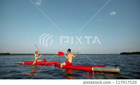 Happy and relaxing young people, man and woman enjoying sunny evening on river, sailing on sup boards with paddles 117017520