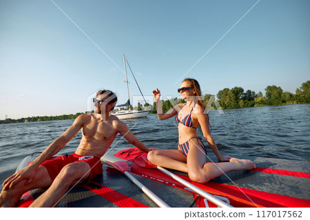Young people, man and woman in swimwear and sunglasses enjoying sunny day on sup boards on river. Paddle boarding activity Young people, man and woman in swimwear and sunglasses enjoying sunny day on sup boards on river. Paddle boarding activity 117017562