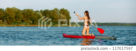 Young woman in swimsuit paddleboarding on sunny day, kneeling on sup board and paddling confidently. 117017597