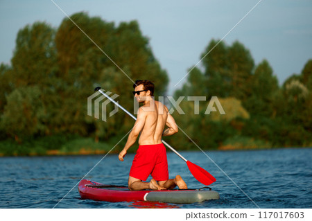 Muscular shirtless man in red swim shorts enjoying summer day while sailing on sup board with paddle, green bank on background. Muscular shirtless man in red swim shorts enjoying summer day while sailing on sup board with paddle, green bank on background. 117017603