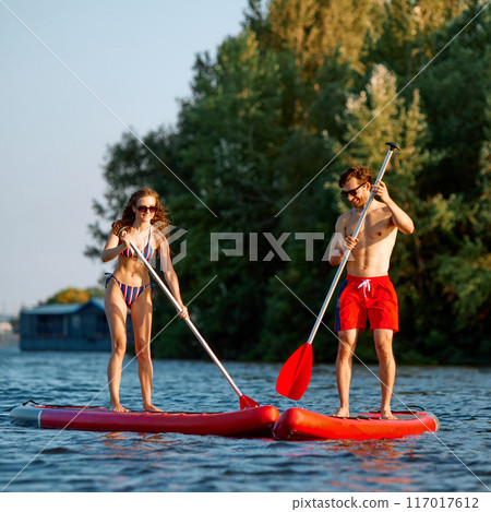 Essence of summer fun. Young man and woman in swimwear standing on paddle board in the middle of the river, with greenery on background Essence of summer fun. Young man and woman in swimwear standing on paddle board in the middle of the river, with greenery on background 117017612