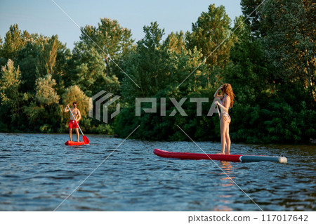 Active weekends. Young man and woman in swimwear enjoying summer day around nature, paddle boarding under clear blue sky. 117017642