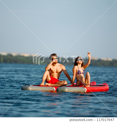 Young man and woman in swimwear and sunglasses sitting in the middle of the river on paddle boards and enjoying view 117017647