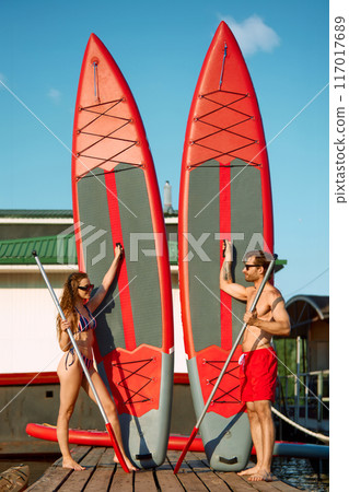 Fit couple, man and woman standing next boards and paddles, preparing for paddleboarding on sunny summer day Fit couple, man and woman standing next boards and paddles, preparing for paddleboarding on sunny summer day 117017689