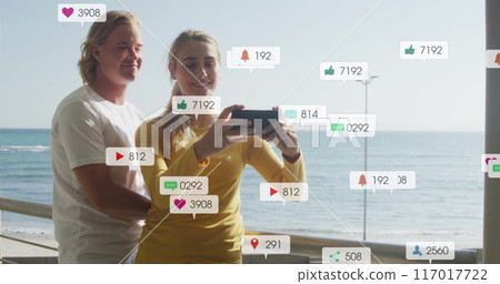 Social media notifications image over couple taking selfie by sea 117017722