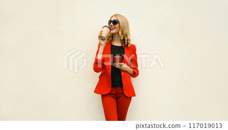 Stylish businesswoman drinking coffee, beautiful woman using smartphone in red business suit 117019013
