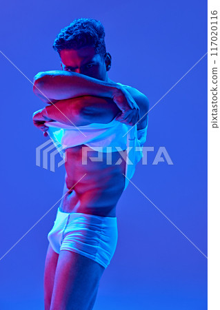 Muscular young man in white underwear lifts his shirt, revealing his chiseled abs in neon light against deep blue background. 117020116