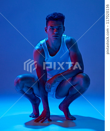 Fit young man in white tank top and underwear crouches low and confidently looks at camera in neon light against deep blue background. Fit young man in white tank top and underwear crouches low and confidently looks at camera in neon light against deep blue background. 117020136