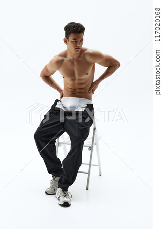 Fit young man sits on chair, shirtless, in black pants and white sneakers, with his hands on his hips against white studio background. 117020168