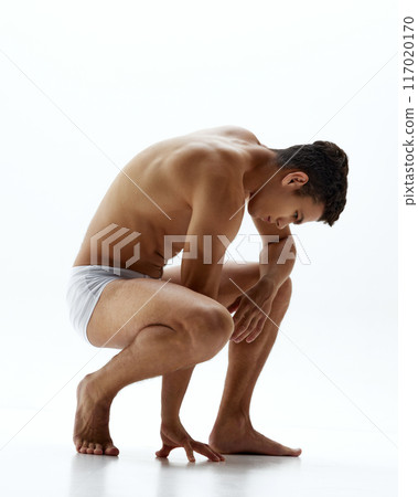 Muscular young man in white boxer briefs crouches low, one hand touching the floor against white studio background. 117020170