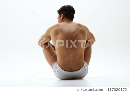 Rear view photo of fit young man in white boxer briefs sits on floor shows his healthy back, spine against white background. 117020173