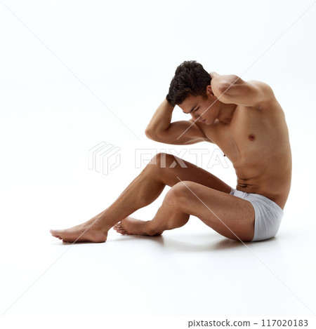 Fit young man in boxer briefs sits on floor with his hands behind his head, showing off his toned muscles against white studio background. 117020183