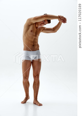 Young man in white boxer briefs stretching his back raising hands an doing bends against white studio background. 117020189