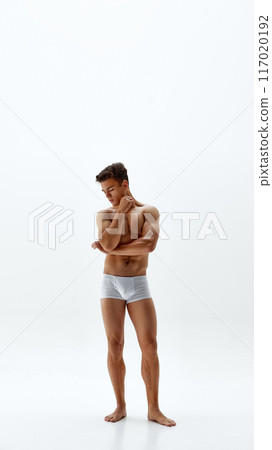 Fit young man in white boxer briefs stands in contemplative pose, hand on chin and arm crossed against white studio background. 117020192