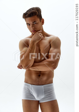 Young naked man posing in boxers gazes at camera with confident expression against white studio background. Young naked man posing in boxers gazes at camera with confident expression against white studio background. 117020193