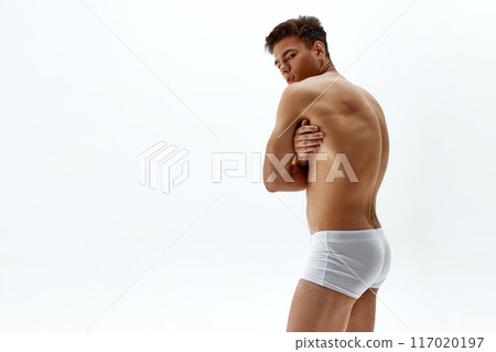 Muscular young man in white boxer looks over shoulder, his toned back and arms crossed against white studio background. Muscular young man in white boxer looks over shoulder, his toned back and arms crossed against white studio background. 117020197