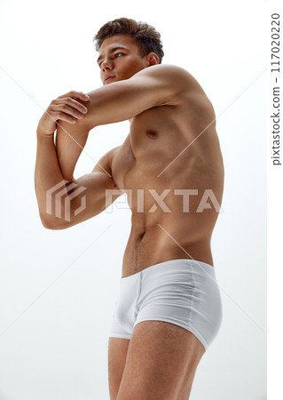 Fit young man in white boxer briefs stretches his arm, displaying his muscular and toned physique against white studio background. Fit young man in white boxer briefs stretches his arm, displaying his muscular and toned physique against white studio background. 117020220