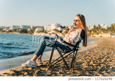 Young beautiful woman in cozy sweater and sunglasses enjoying hot coffee from thermo mug while relaxing on winter seaside sand beach. Cute attractive girl drinks tea taking sunbathe near ocean 117020246