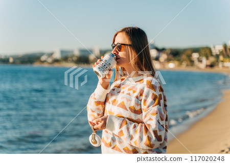 Young beautiful woman in cozy sweater and sunglasses enjoying hot coffee from thermo mug while relaxing on winter seaside sand beach. Cute attractive girl drinks tea taking sunbathe near ocean Young beautiful woman in cozy sweater and sunglasses enjoying hot coffee from thermo mug while relaxing on winter seaside sand beach. Cute attractive girl drinks tea taking sunbathe near ocean 117020248