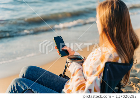 Young beautiful woman holding texting smartphone cellphone while drinking coffee on winter beach with sea waves in background. Cute attractive girl using mobile when enjoying tea on autumn seashore Young beautiful woman holding texting smartphone cellphone while drinking coffee on winter beach with sea waves in background. Cute attractive girl using mobile when enjoying tea on autumn seashore 117020282